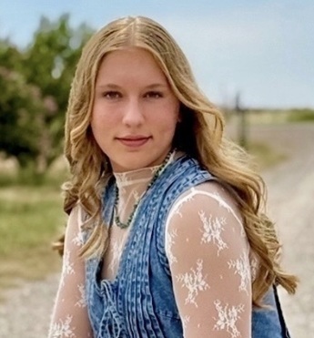 A teen girl in a lace shirt and denim vest poses for a photo in an outdoor setting.