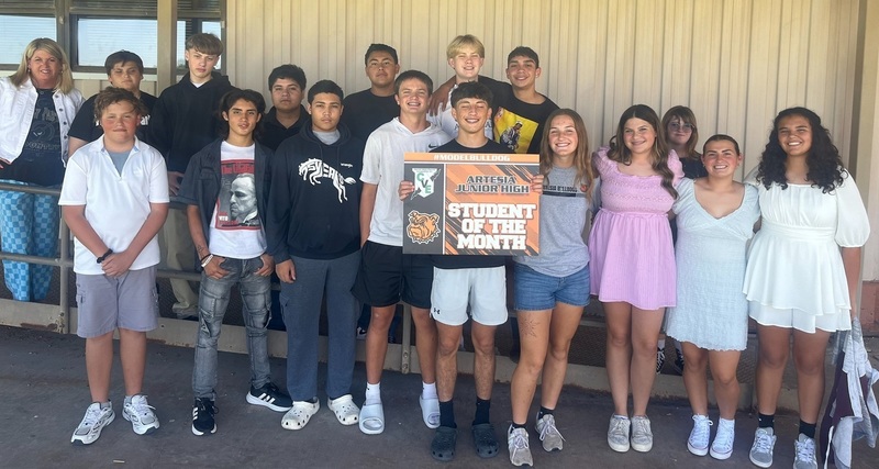 A teen boy in a black t-shirt and white shorts holds a sign reading "Student of the Month", surrounded by classmates and a female teacher.