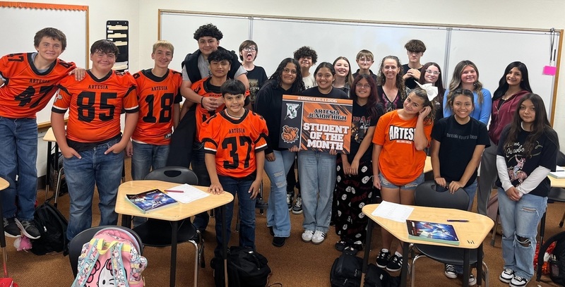 A teen girl in a black t-shirt and jeans holds a sign reading "Student of the Month", surrounded by classmates and a female teacher.
