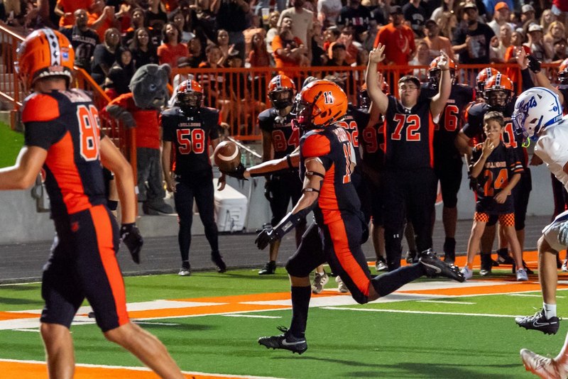 A teen boy in a black-and-orange football uniform runs toward the end zone with the ball outstretched in his right hand as teammates on the sideline and the home crowd look on.