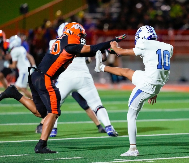 A teen boy in a black-and-orange football uniform lungs with his arms in front of him to block the football as an opponent in white and blue attempts to punt it.