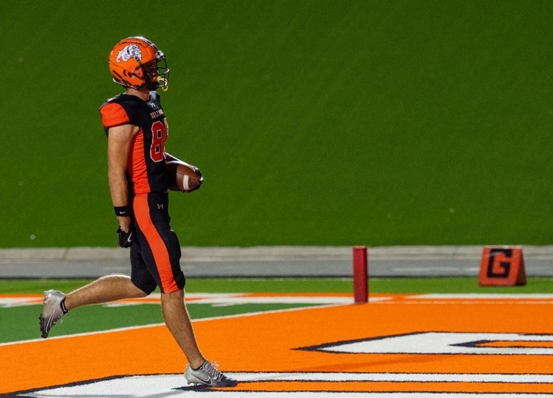 A teen boy in a black-and-orange football uniform walks into the end zone untouched.
