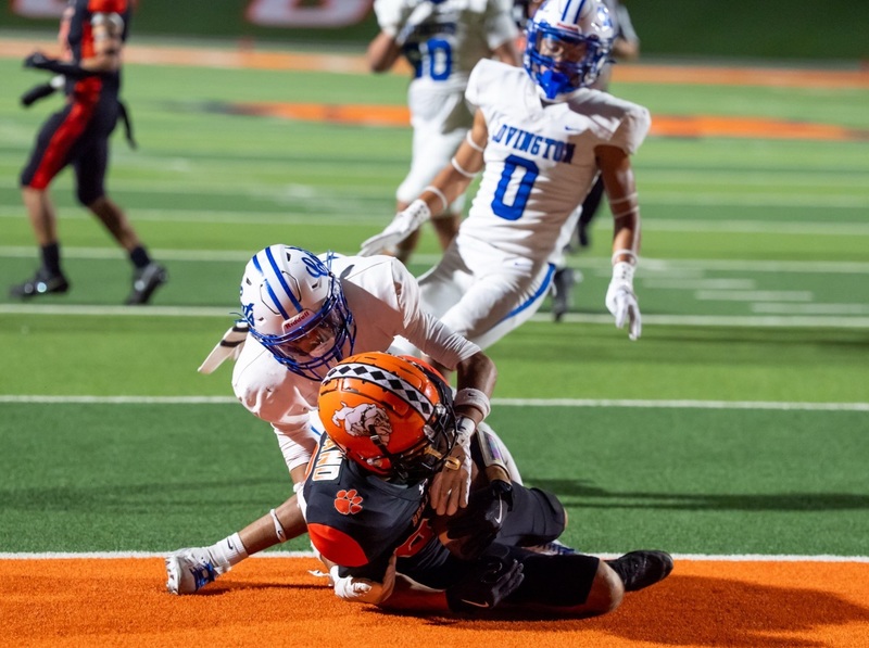 A teen boy in a black-and-orange football uniform falls into the end zone as he's tackled from behind by an opponent in white and blue.