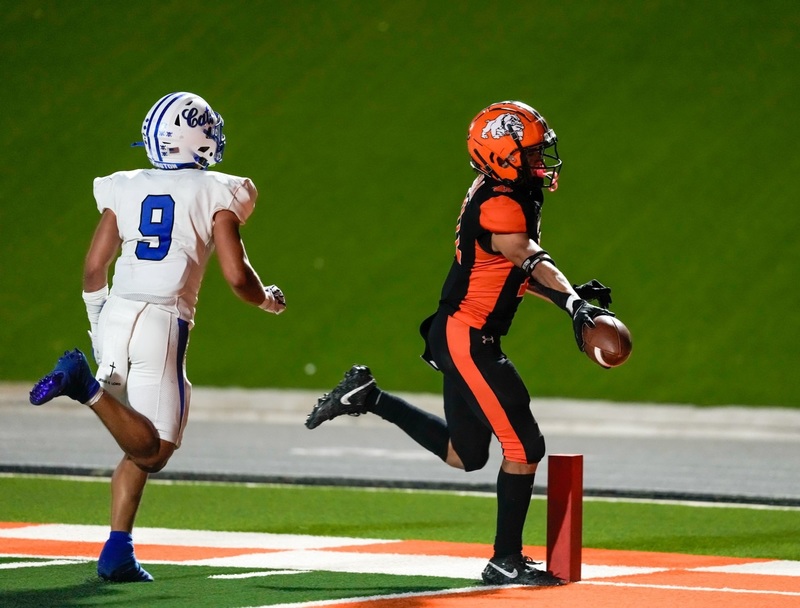 A teen boy in a black-and-orange football uniform runs past the pylon with the ball outstretched in his right hand as an opponent in white and blue attempts to catch up.