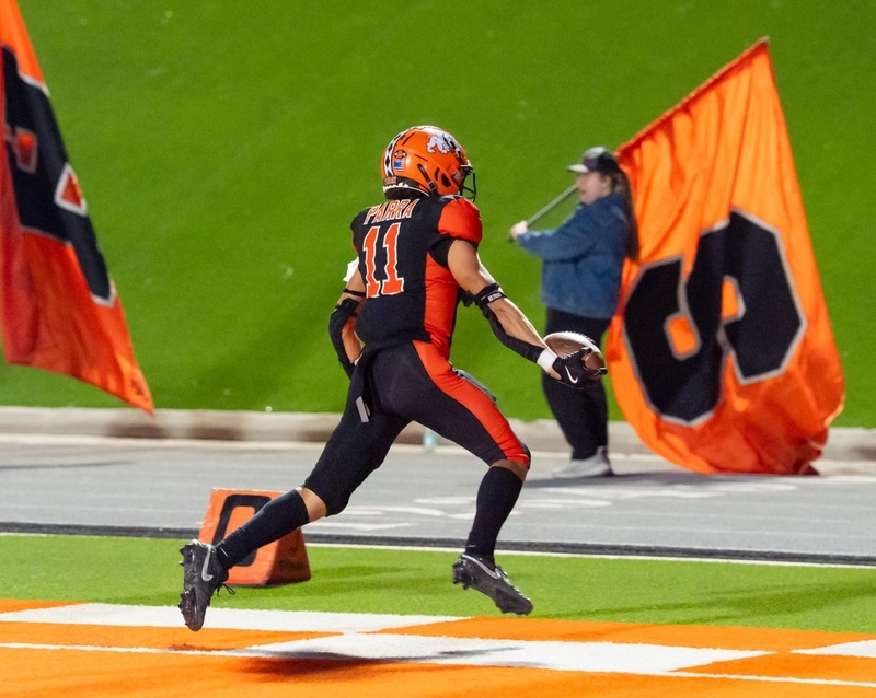 A teen boy in a black-and-orange football uniform runs across an orange-and-white checkerboard end zone with the ball in his right hand as large orange flags bearing the letters "H" and "S" are waved in the background.