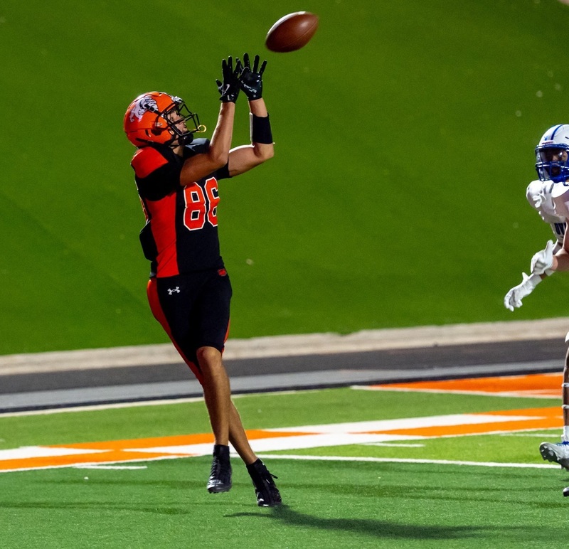 A teen boy in a black-and-orange football uniform raises his hands and looks at the football as it flies toward him.