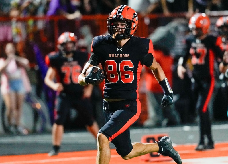 A teen boy in a black-and-orange football uniform runs with the ball in his right hand.