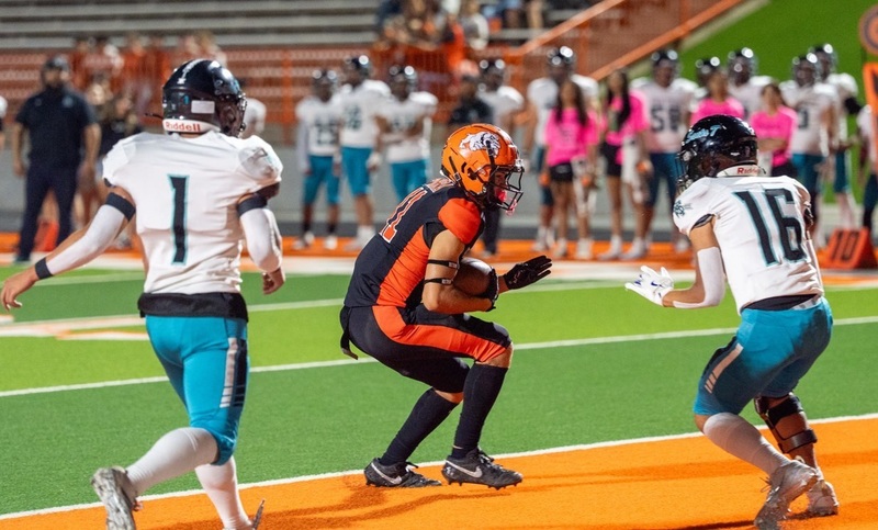 A teen boy in a black-and-orange football uniform hunches down as he runs into the end zone with the ball in his right elbow as two opponents in white and teal look on.