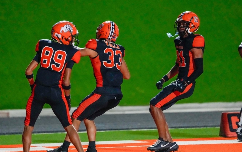 A teen boy in a black-and-orange football uniform celebrates in the end zone with two teammates.