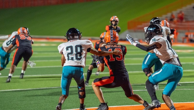 A teen boy in a black-and-orange football uniform catches the ball with his back to the camera as two opponents in white and teal move in to tackle him.
