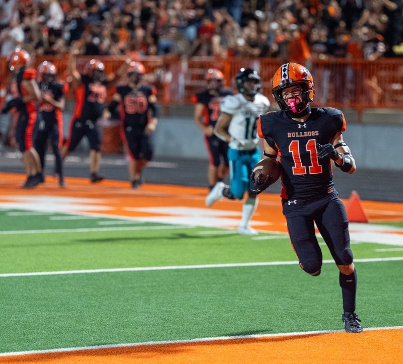 A teen boy in a black-and-orange football uniform runs over the goal line with the ball in his right hand as teammates and the home crowd cheer in the background.