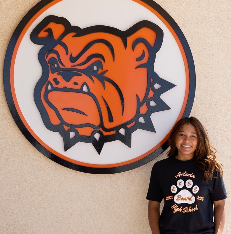 A teen girl in a black t-shirt with a white paw print on the front poses next to a Bulldog insignia.
