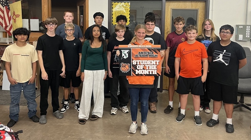 A teen girl in an orange hoodie and black leggings holds a sign reading "Student of the Month", surrounded by classmates.