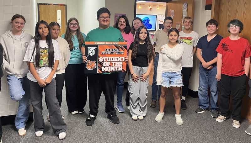 A teen boy in a teal pullover and black pants holds a sign reading "Student of the Month", surrounded by classmates.