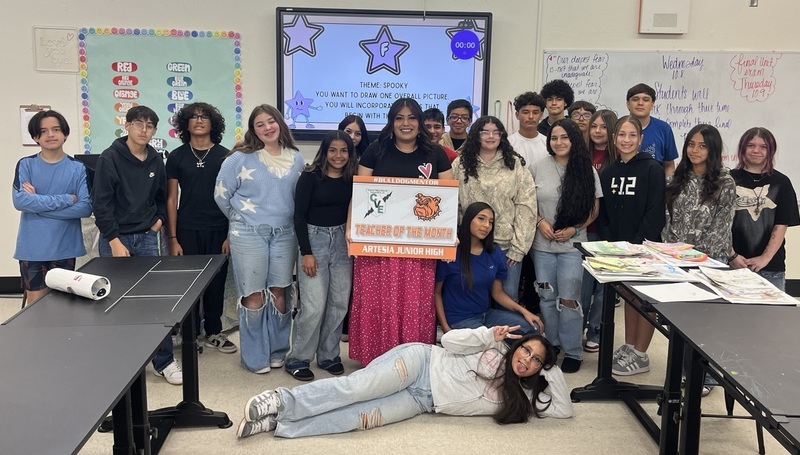 A female teacher in a black shirt and pink skirt holds a sign reading "Teacher of the Month", surrounded by students.