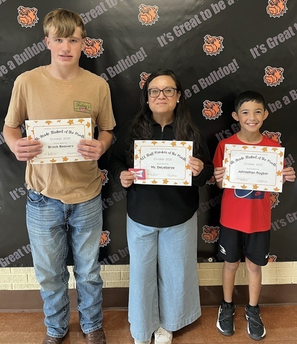 A preteen boy in a tan shirt and jeans, a female teacher in a black shirt and jeans, and a preteen boy in a red shirt and black shorts stand holding certificates.