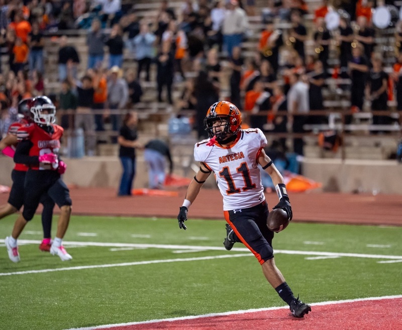 A teen boy in a white-and-orange football uniform bearing the number 11 steps over the goal line while holding the football in his left hand.