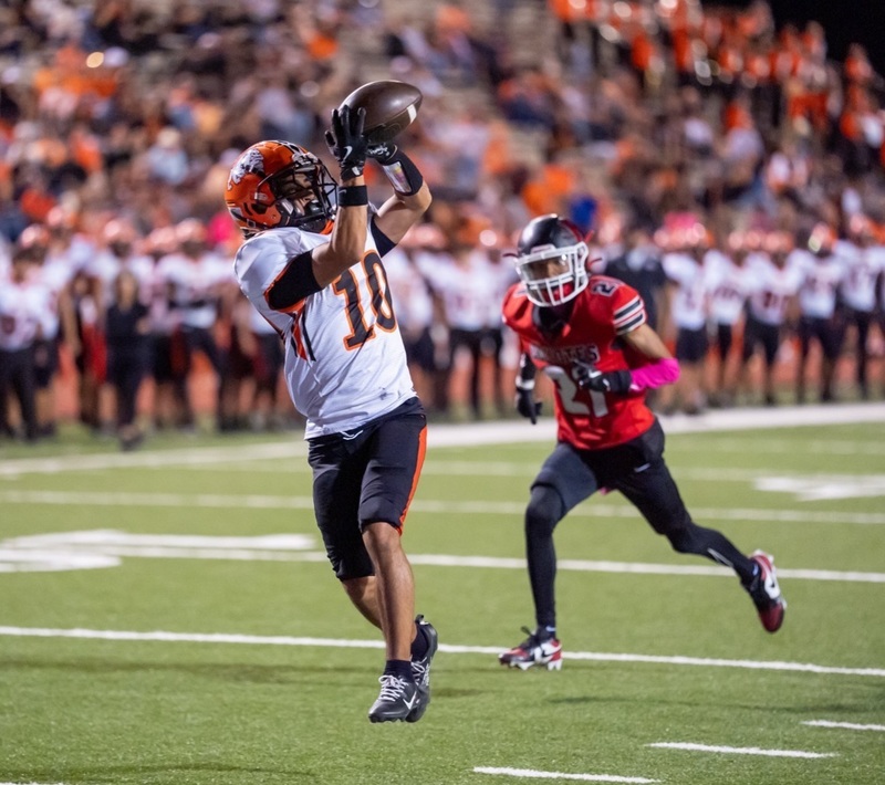 A teen boy in a white-and-orange football uniform looks the football into his hands, which are raised to eye level, as an opponent in red and black chases after him.