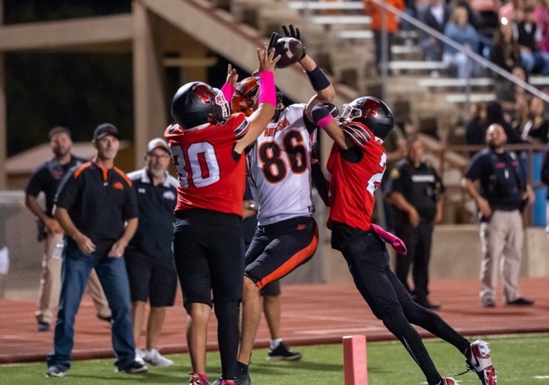 A teen boy in a white-and-orange football uniform bearing the number 86 leaps up to catch the football with opponents in red and black on both of his sides.