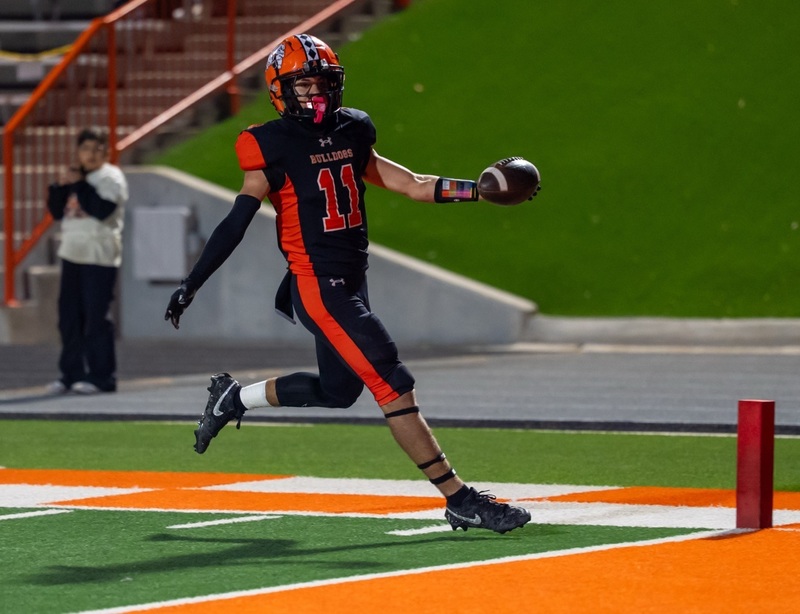 A teen boy in a black-and-orange football uniform extends the ball in his left hand as he runs over the goal line.