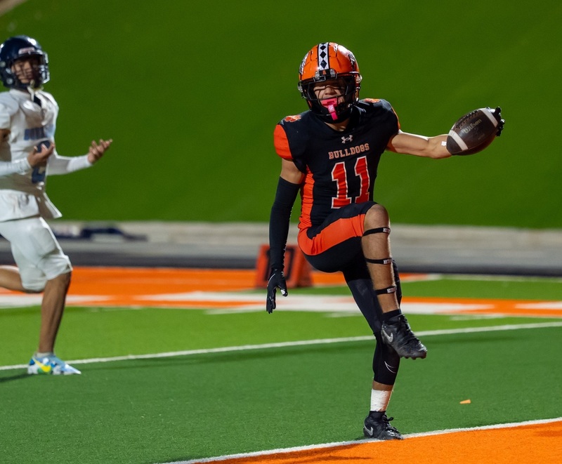 A teen boy in a black-and-orange football uniform high-steps across the goal line with the ball in his left hand ahead of an opponent in white and baby blue.