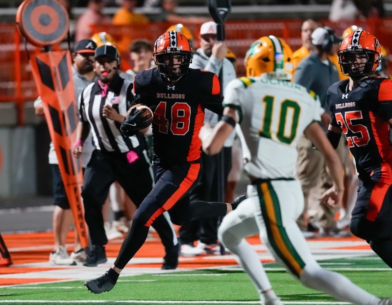 A teen boy in a black-and-orange football uniform carries the ball in his right arm as he looks at an opponent in white, gold and green.