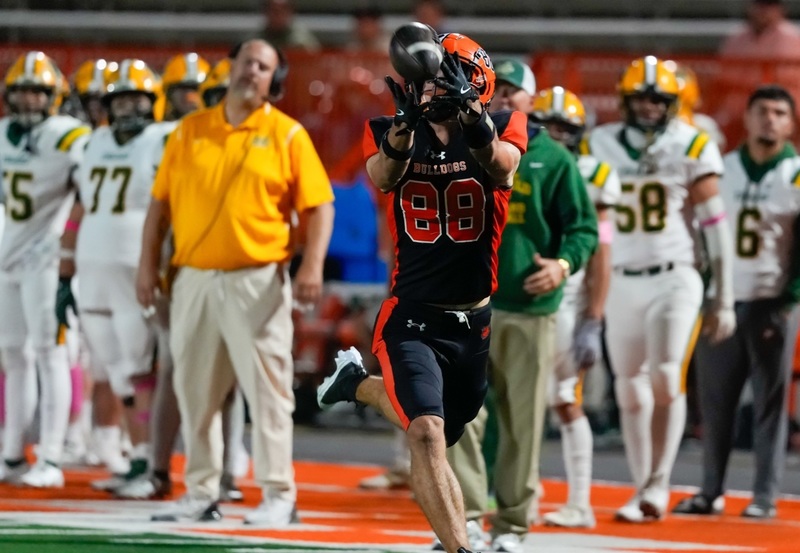 A teen boy in a black-and-orange football uniform lunges forward to catch the football in his fingertips.