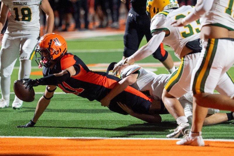 A teen boy in a black-and-orange football uniform dives forward with the ball extended in his left hand as an opponent in white, gold and green attempts to drag him down from his waist.