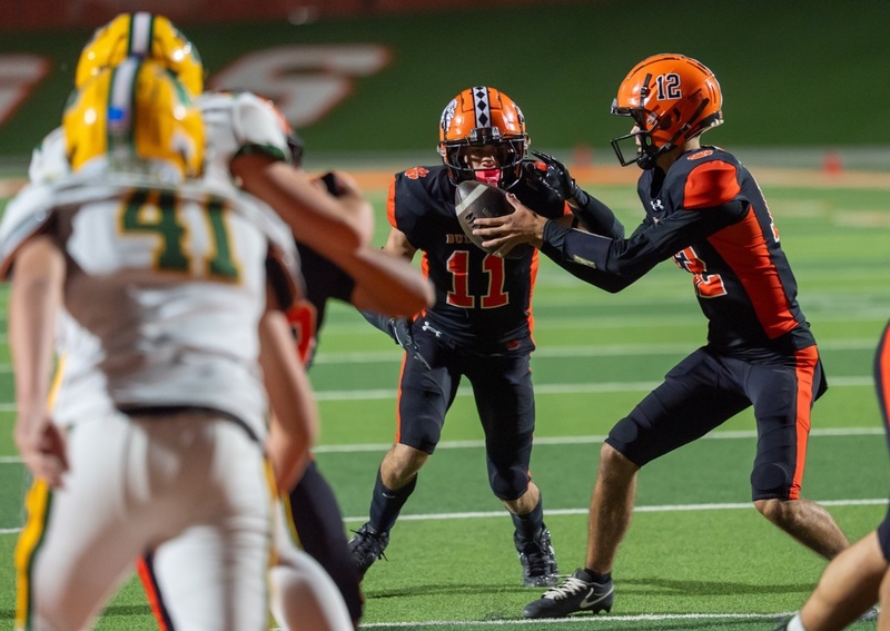 A teen boy in a black-and-orange football uniform hands the football to a teammate as opponents in white, gold and green rush them.
