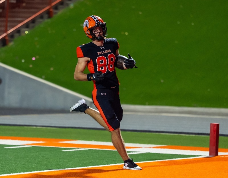 A teen boy in a black-and-orange football uniform bearing the number 88 steps into an orange end zone with the ball tucked into his left elbow.