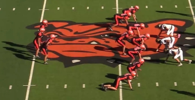 An offensive unit in black-and-orange football uniforms lines up against opponents in white, black and gold in a T formation on a football field with a large graphic of a Bulldog in its center.