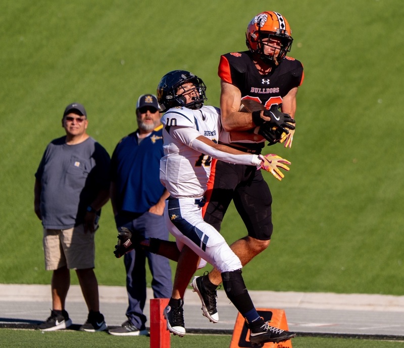 A teen boy in a black-and-orange football uniform jumps into the air to catch a ball over an opponent in white, black and gold.