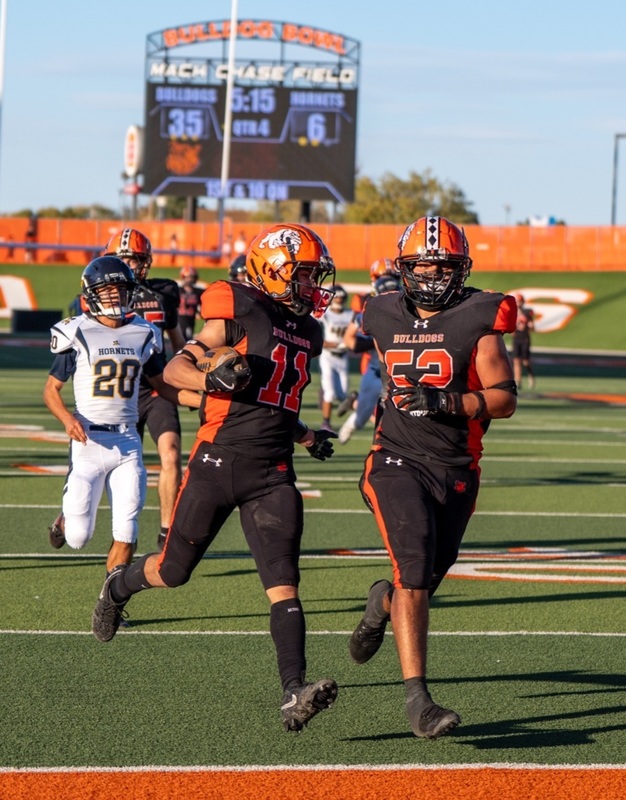 A teen boy in a black-and-orange football uniform holding a football in his right elbow follows a teammate into the end zone.