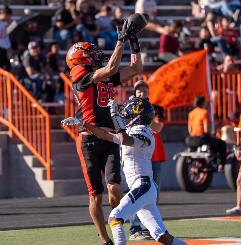 A teen boy in a black-and-orange football uniform jumps in the air as a football sails into his hands over an opponent in white, black and gold.