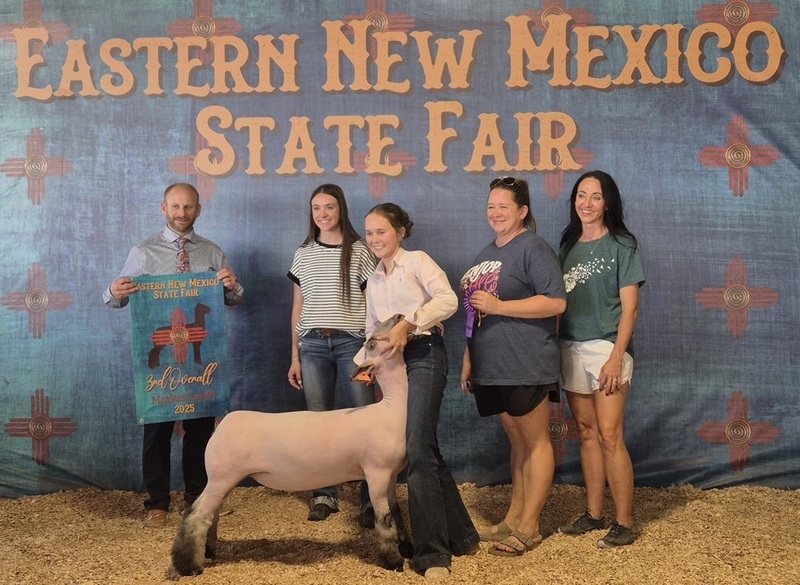 A teen girl stands with a white-and-black lamb, surrounded by three other women and a male holding a banner.