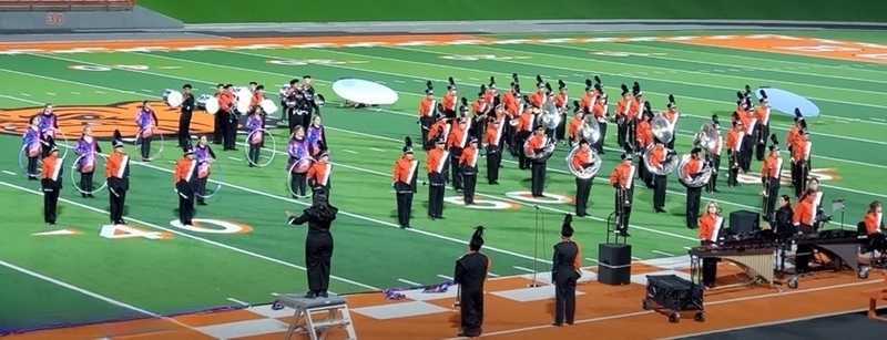 A high school band in orange, black and white uniforms stands at attention on a football field.
