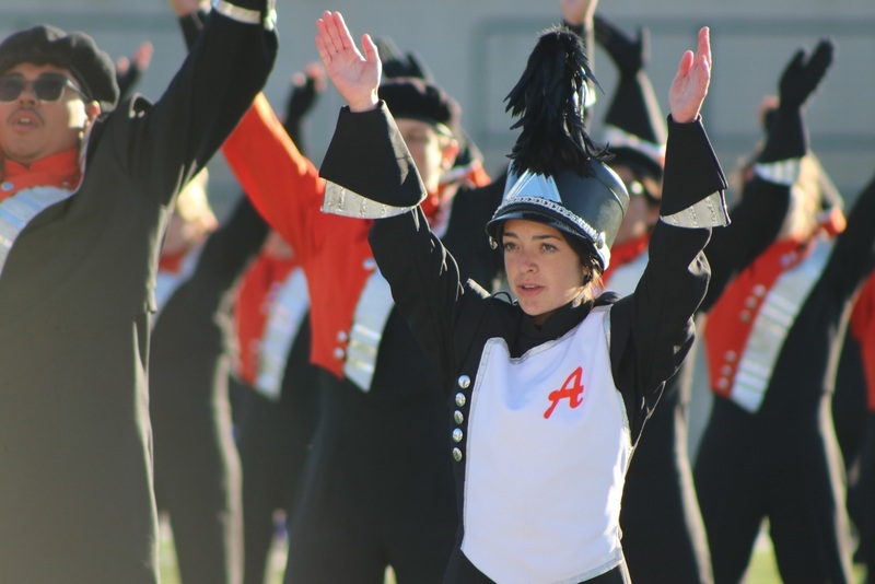 Male and female teens in orange, black and white band uniforms raise their arms into the air.