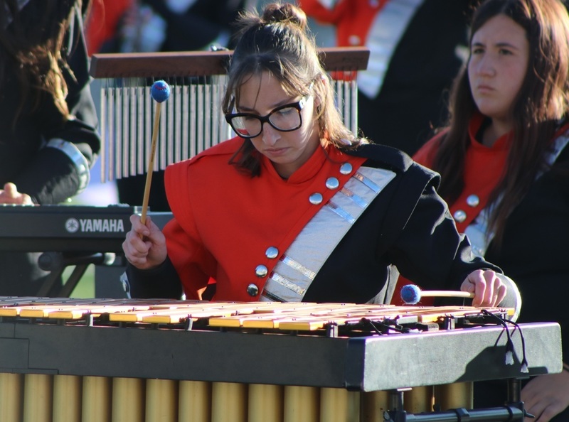 A female teen in glasses and an orange, black and white band uniform plays a marimba.