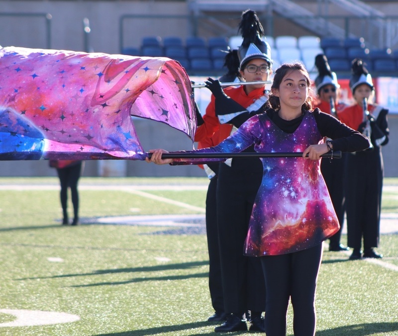 A female teen in a uniform with a galactic print waves a similarly colored flag as band members play flutes behind her.