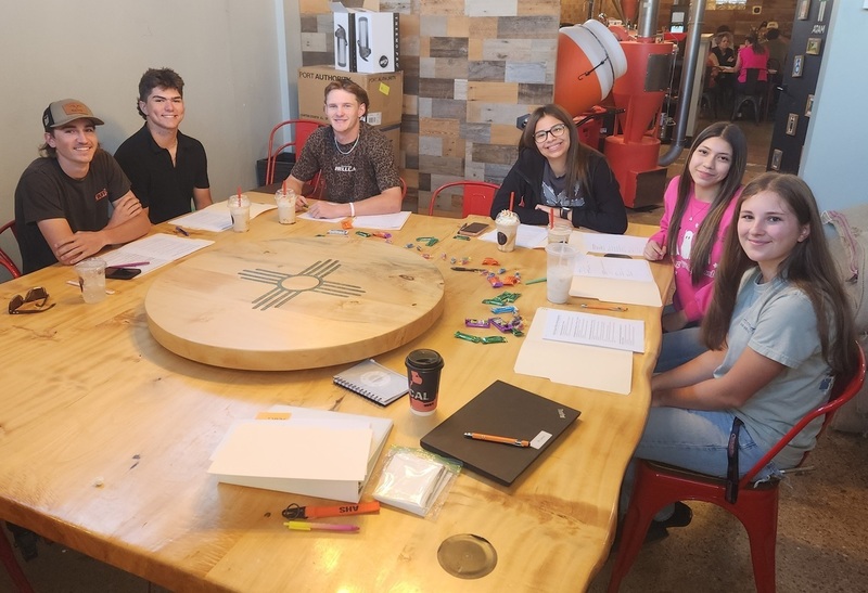 Three male and three female teens smile for the camera while seated around a large wooden table.