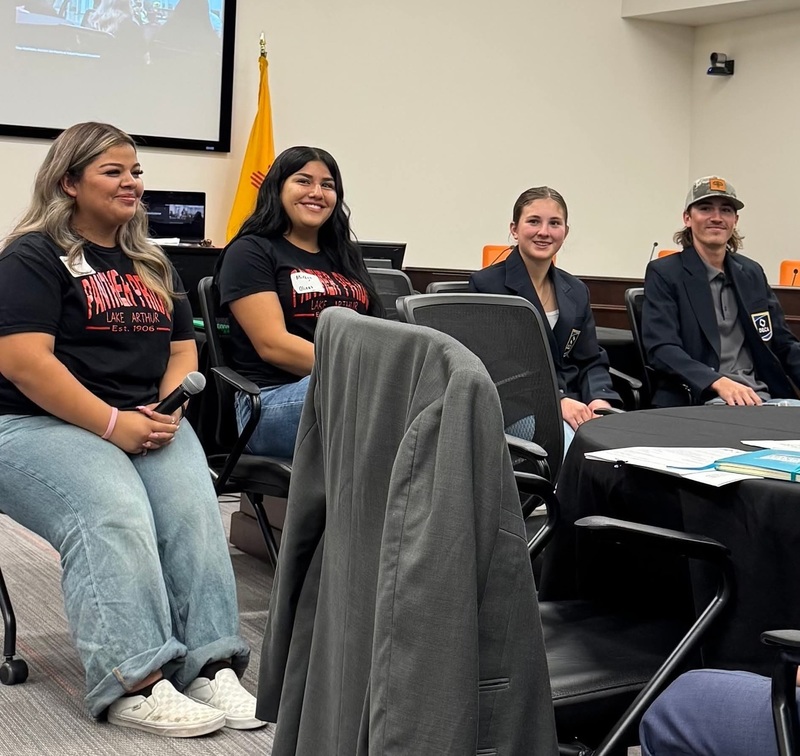 Three female and one male teen smile for the camera while seated in chairs in a large conference room.