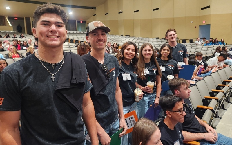 Three male and three female teens stand among other seated teens in an auditorium and smile for the camera.
