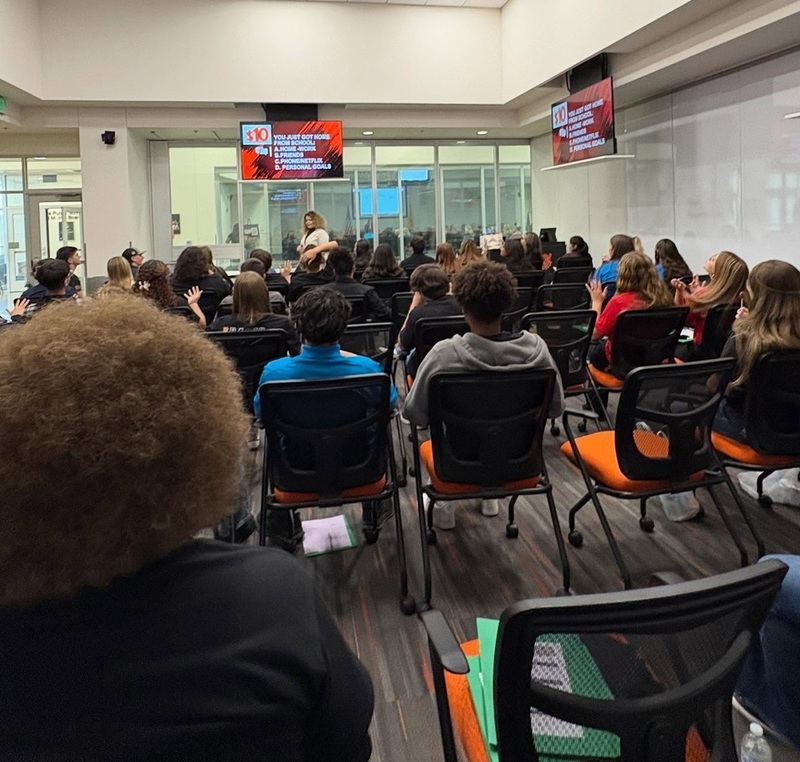 A large group of male and female teens are seen from the back as they sit in chairs in a large conference room.