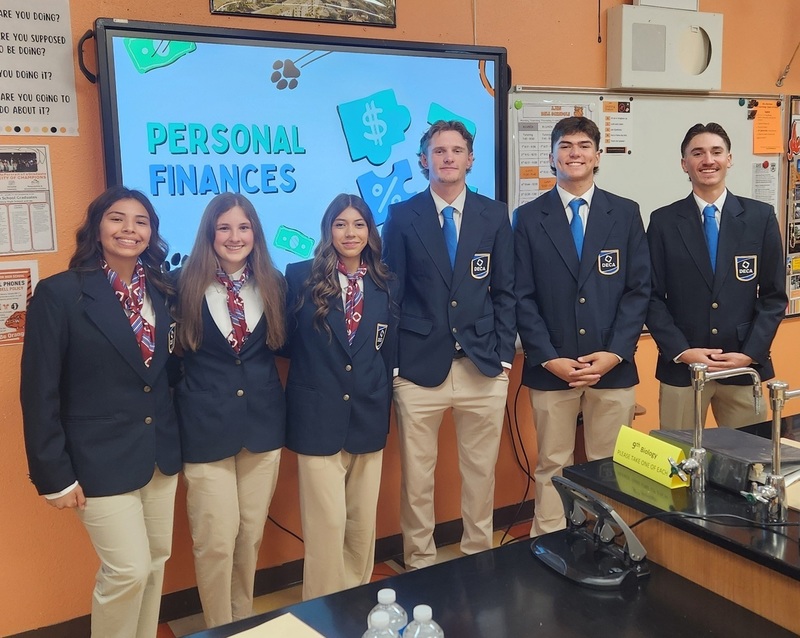 Three female and three male teens in black suit jackets and khaki pants pose for a photo in front of a large screen reading "Personal Finances."