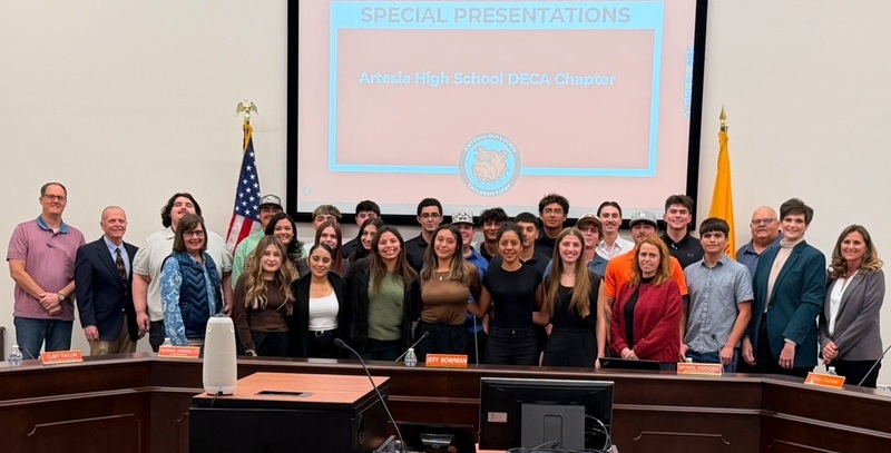 Multiple male and female teens are flanked by men and women as they pose for a photo behind a long desk.