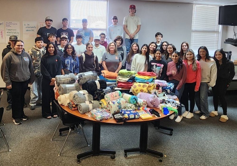 A large group of male and female teens poses for a photo behind a round table laden with blankets and socks.