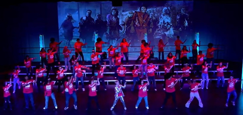 A group of male and female teens in red shirts and jeans does the "Thriller" dance in front of a large screen bearing an image of Michael Jackson.
