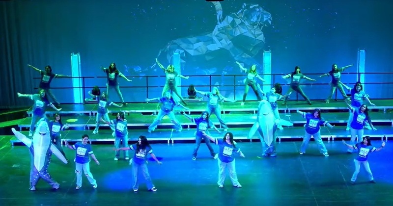 A group of female teens and two students in shark costumes waves their arms as they sing.