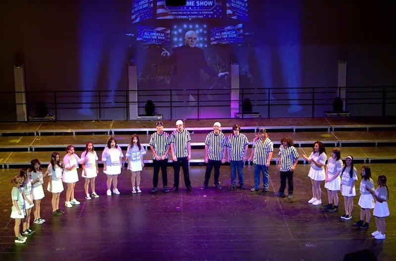 Female and male teens dressed as cheerleaders and referees form a semicircle on a stage.