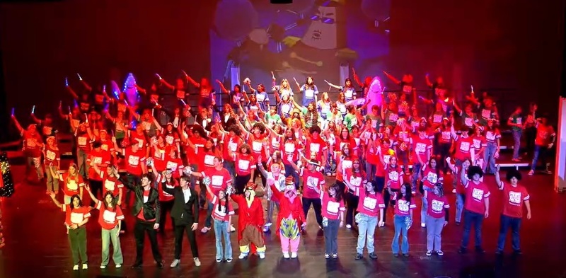 A large group of male and female teens, most in red shirts and two dressed as SpongeBob SquarePants and Patrick Starfish, wave an arm in the air as they sing.